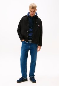 Νέος άνδρας με μαύρο μπουφάν, μπλε καφέ φούτερ, μπλε τζιν και σκούρα αθλητικά παπούτσια, στέκεται κοιτάζοντας προς τα κάτω με το ένα χέρι στην τσέπη, σε λευκό φόντο.