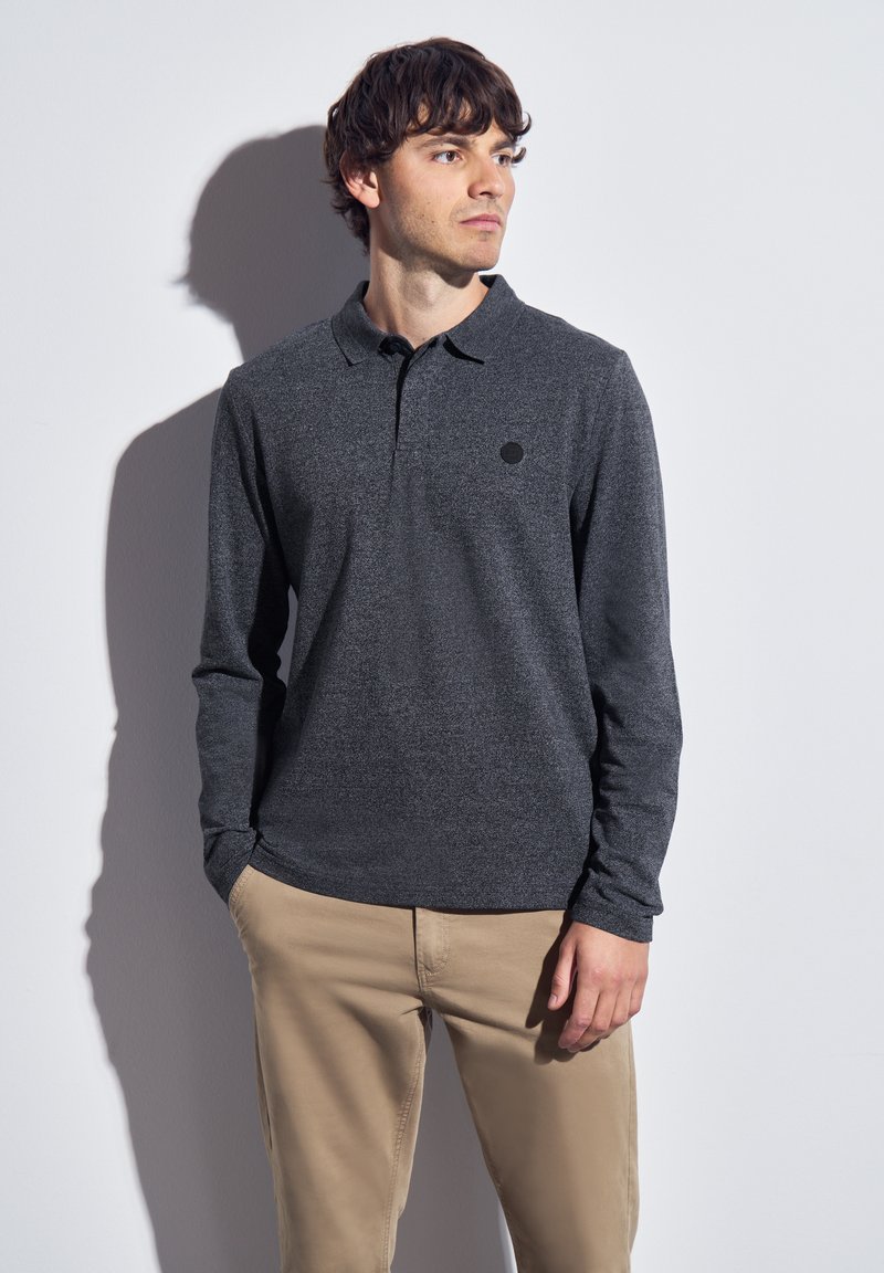 Muž v tmavě šedém polo tričku s dlouhým rukávem a béžových kalhotách stojí s rukama v kapsách, hledí doprava proti jednoduché bílé zdi.