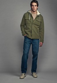 Oliven grøn jakke med tekstureret overflade, to brystlommer og fleeceforet hætte, båret over en beige skjorte og blå jeans. Beige sneakers.