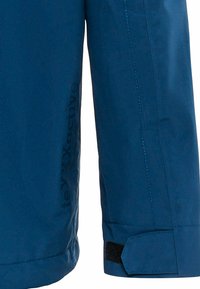 Ärmel einer blauen wasserdichten Jacke mit strukturiertem Stoff, kontrastierenden Nähten und einem Klettverschluss. Logo an der Seite sichtbar.