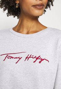 Šedá mikina s kulatým výstřihem, s vyraženým červeným nápisem "Tommy Hilfiger". Měkká textura, středně těžká tkanina, neformální design.