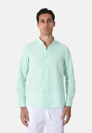 Uomo che indossa una camicia verde chiaro abbottonata con collo alla coreana e pantaloni bianchi, in piedi su uno sfondo chiaro uniforme.