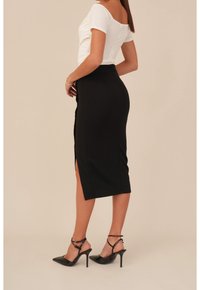 Svart blyertskjol med sidoklyftor, kombinerad med en vit åtsittande topp. Modellen har på sig svarta stilettklackar med ankelremmar. Slät tygtextur.