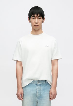 Tricou alb din bumbac cu mâneci scurte, având o broderie subtilă neagră cu textul „Closed” pe partea stângă. Purtați cu blugi de culoare bleu deschis.