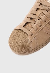 Tan suedesneaker, mille tekstuuriga kummine varvas, mis sisaldab diamantruutude mustrit ja klassikalist nööri süsteemi. Küljel on kerge õmbluste aktsent.
