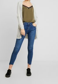 Světle šedý dlouhý kardigan, olivová saténová tílko s černou krajkou, modré skinny džíny se třepenými lemy a černé šněrovací tenisky.