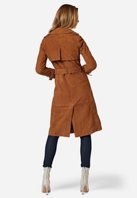 Brown suede trench coat with a belted waist, back vent, and wide collar. Paired with dark blue jeans and white high-heeled boots.
