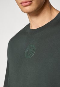 Mørkegrøn kortærmet t-shirt lavet af bomuld, med et tekstureret rundt mærke med "Chelsea Football Club" logoet på brystet.