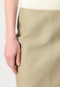 Beige skinnskjørt med glatt tekstur, med en tettsittende design og rett silhuett. Hånd som hviler på siden, som viser naturlige negler.