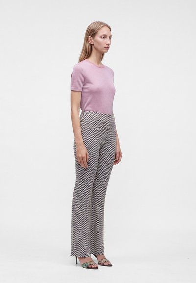 Femme debout, face à droite, portant un haut rose à manches courtes, un pantalon taille haute à motif chevron et des sandales à talons ouverts.