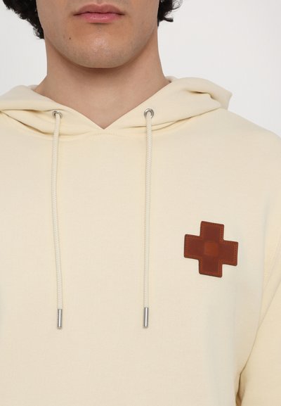 Cream hoodie with a textured brown leather patch featuring a cross design. Includes a drawstring hood and silver-tone hardware.