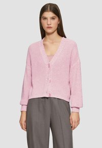Rosa gestrickte Strickjacke mit V-Ausschnitt, vier Knöpfen und überschnittenen Schultern, kombiniert mit grauen weiten Hosen. Strukturierter Stoff mit gerippten Bündchen.