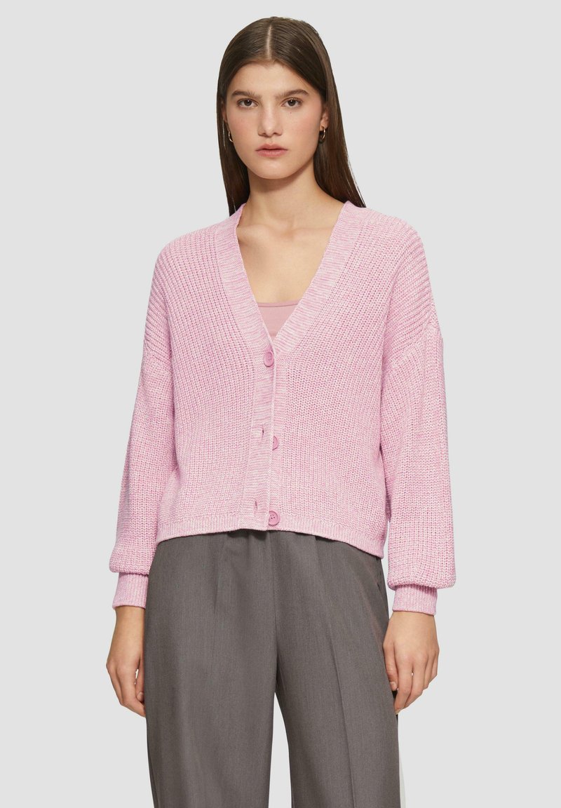 Rosa gestrickte Strickjacke mit V-Ausschnitt, vier Knöpfen und überschnittenen Schultern, kombiniert mit grauen weiten Hosen. Strukturierter Stoff mit gerippten Bündchen.