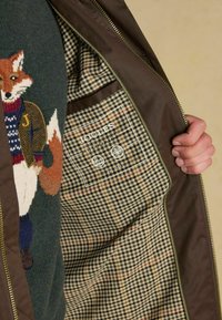 Brązowa kurtka z widocznym swetrem w zielony i lisie wzory. Podszewka w kratkę z zielonymi, beżowymi i czerwonymi akcentami. Wyraźna metka Joules.