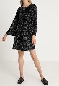 Vestido negro con patrones abstractos blancos, mangas acampanadas largas, cuello redondo y un corte holgado, combinado con zapatos negros tipo slip-on.