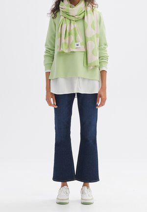 Person trägt hellgrünen Pullover über weißem Hemd, grüner und beiger gemusterter Schal, dunkle ausgestellte Jeans und weiße Sneaker und steht.