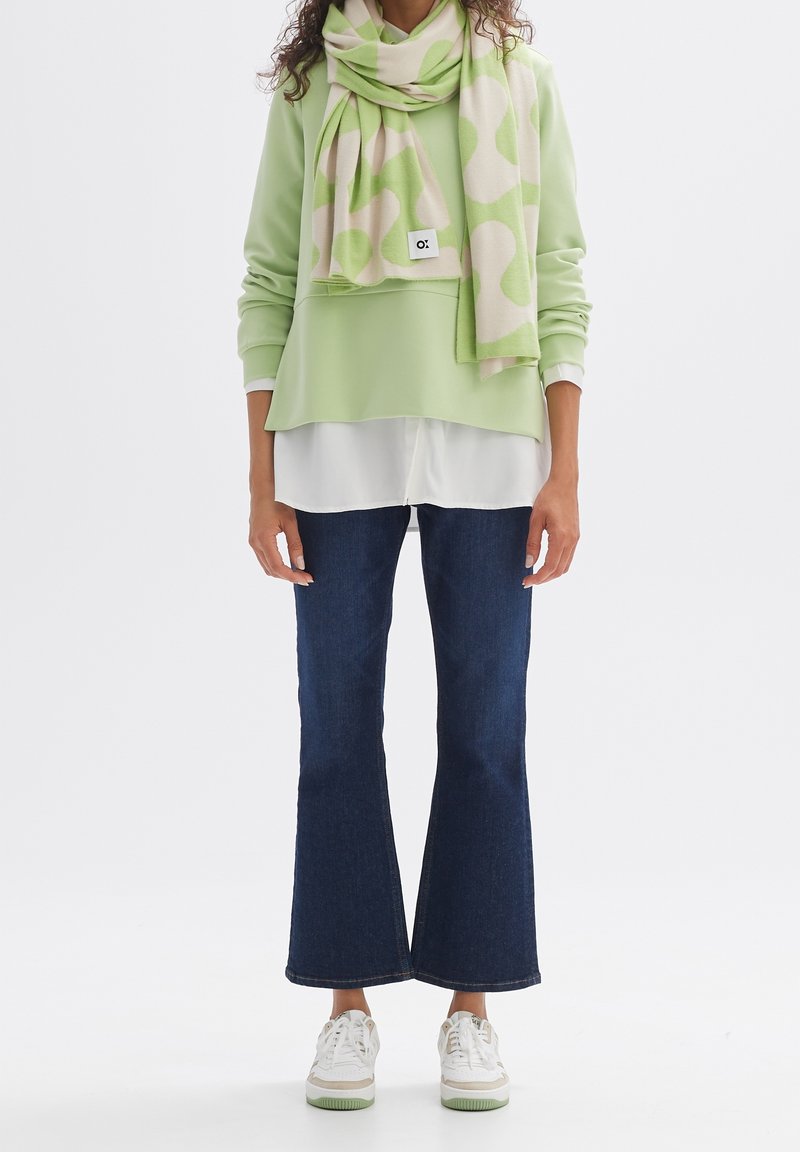 Personne portant un pull vert clair superposé à une chemise blanche, une écharpe à motifs verts et beiges, un jean évasé foncé et des baskets blanches, debout.