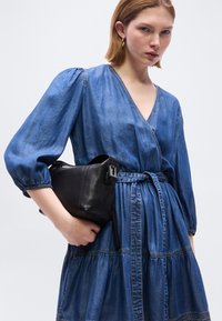 Γυναίκα που φοράει μπλε φόρεμα τύπου ντενίμ με φουσκωτά μανίκια, κρατώντας μια μαύρη δερμάτινη τσάντα χειρός σε ουδέτερο φόντο.