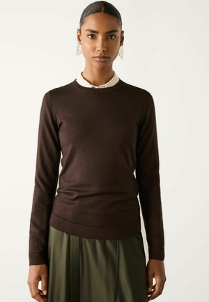 Kvinde iført en mørkebrun langærmet sweater over en hvid skjorte med krave og et olivengrønt skørt, der vender fremad med et neutralt udtryk.