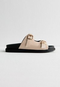 Sandalia de deslizamiento sintética beige con acentos de doble hebilla dorada y suela negra. Diseño plano con textura suave. Sin elevación del tacón.