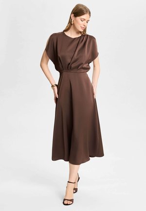 Femme portant une robe midi marron avec des manches courtes et une taille plissée, accessoirisée avec des boucles d'oreilles dorées, un bracelet et des talons noirs à bride cheville.