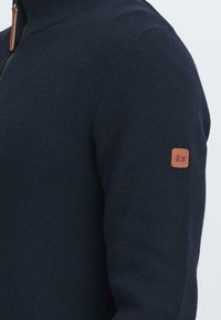 Suéter de cremallera azul marino en tejido de punto texturizado; presenta un cuello alto y un parche de logo de cuero marrón en la manga izquierda.