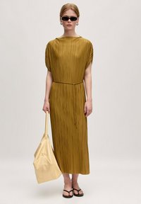 Faltenrock Kleid in Senfgelb mit kurzen Ärmeln, einem gebundenen Taillengürtel und einem fließenden Design. Kombiniert mit einer cremefarbenen Tasche und schwarzen Sandalen. Sonnenbrille getragen.