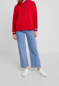 Felpa rossa in cotone con cappuccio regolabile, caratterizzata da dettagli blu e polsini a righe. Abbinata a jeans a gamba larga di un blu chiaro e scarpe da ginnastica bianche.
