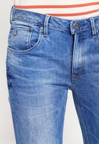 Los jeans de mezclilla en un tono azul claro cuentan con un diseño de cinco bolsillos, con cremallera y cierre de botón. El tejido tiene una textura ligeramente desgastada.
