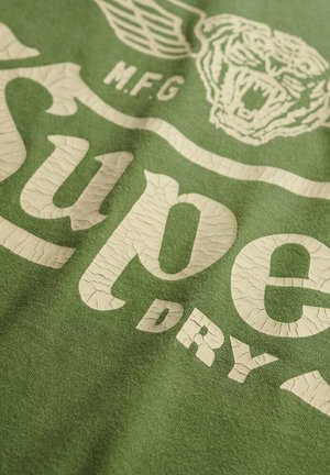 Camiseta de algodón verde con letras blancas desgastadas "Super Dry" y un gráfico de tigre. Acabado texturizado con elementos de diseño de estilo vintage.
