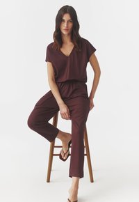 Bordeaux jumpsuit met korte mouwen, V-hals, elastische tailleband en taps toelopende benen. Draag het met bruine slippers terwijl je op een houten kruk zit.