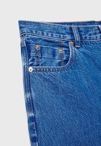 Détail d'un jean en denim bleu montrant une poche avant, une poche monnaie, une passants de ceinture, une double couture et des rivets métalliques sur un fond clair.