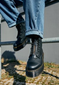 Bottines en cuir noir avec une semelle épaisse, des fermetures éclair en métal et des coutures contrastantes. Portées avec un pantalon en jean bleu retroussé.