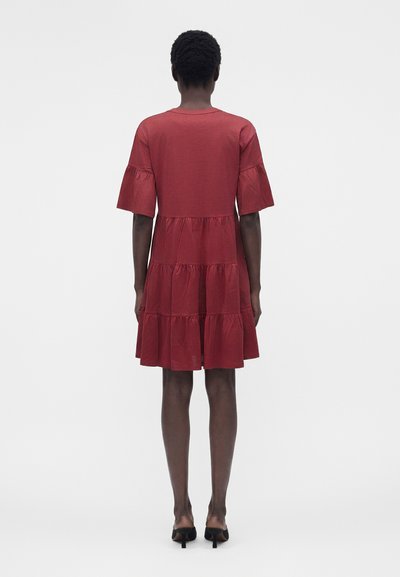 Γυναίκα με κοντά, σγουρά μαλλιά, που φορά κόκκινο φόρεμα με μήκος έως το γόνατο, σε επίπεδα με καμπανοειδή μανίκια και μαύρα παπούτσια με τακούνι, στέκεται γυρισμένη με την πλάτη.