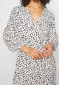 Robe blanche avec un décolleté en V profond, des manches bouffantes et une taille cintrée, ornée d'un motif à pois noirs.