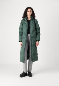 Lang grønn pufferjakke med høy krage, quiltet design og frontglidelås. Kombinert med lysegrå jeans og svarte klumpete sko.