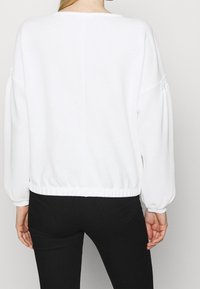 Bílý pletený pulovr s kulatým výstřihem, spadenými rukávy a elastickým lemem. Design doplňuje strukturovaná látka a rukávy připomínající kapsu.