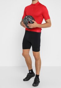 Red short-sleeved cycling jersey with a high collar, paired with black cycling shorts. The model holds a black helmet. Black cycling shoes are worn.