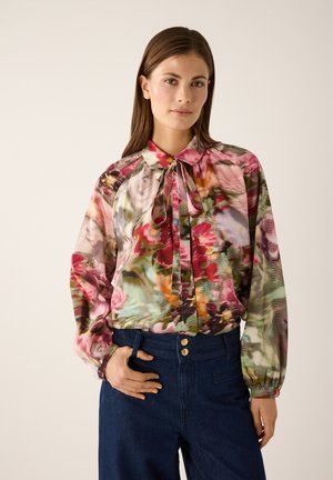 Florale Bluse aus transparentem Stoff mit Schleifenkragen, mehrfarbiges Muster in Pink und Grün, lange Ballonärmel, kombiniert mit dunklen Jeans.