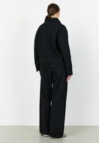 Svart sweatshirt och vida sweatpants i elastiskt material. Noterbara detaljer inkluderar en hög krage och ett präglat logotyp på byxorna.