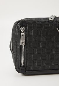 Black synthetic leather bag with embossed geometric pattern, silver hardware, zippers on sides, and logo detail on the front.