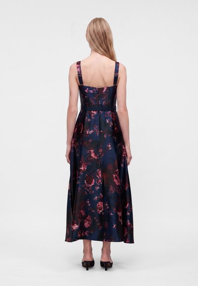 Femme aux longs cheveux blonds portant une robe florale foncée sans manches avec des bretelles ajustables et des talons hauts noirs, debout de dos sur fond blanc.