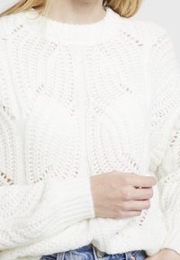 Biała swetrowa bluzka z fakturowym wzorem, okrągłym dekoltem i ściągaczami. Noszona na dżinsach. Wyróżnia się splatanym wzorem i ażurowym splotem.