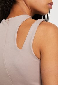 Vaaleanbeige hihaton paita, jossa on pyöreä kaula-aukko, kolmion muotoinen aukko ja piilotettu vetoketju takana. Sileä ja joustava materiaali.