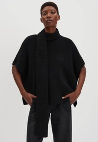 Persona che indossa un poncho nero a maglia a collo alto, largo, con un lungo dettaglio di sciarpa, abbinato a pantaloni neri, mani nelle tasche, sfondo semplice.