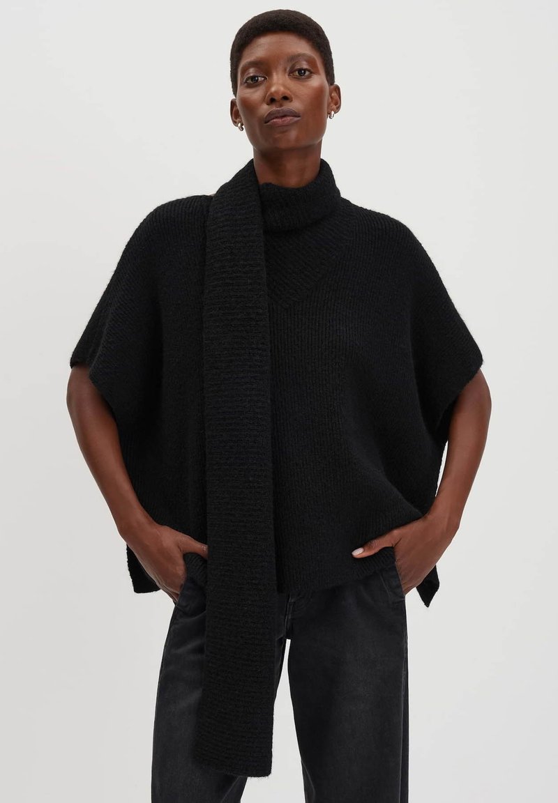Persona che indossa un poncho nero a maglia a collo alto, largo, con un lungo dettaglio di sciarpa, abbinato a pantaloni neri, mani nelle tasche, sfondo semplice.