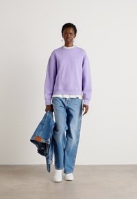 Felpa lavanda, jeans blu larghi, sneakers bianche; tiene una giacca di jeans. Design semplice con un piccolo logo sulla felpa.