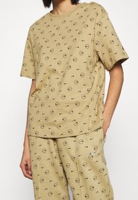 Bavlněné tričko v světle hnědé barvě s černými kulatými grafikami a logy Nike, s uvolněným střihem a krátkými rukávy.