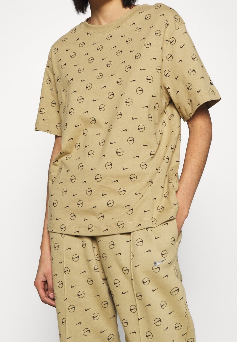 Bavlněné tričko v světle hnědé barvě s černými kulatými grafikami a logy Nike, s uvolněným střihem a krátkými rukávy.