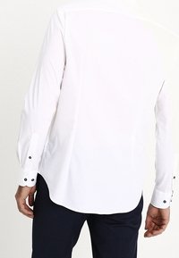 Uomo che indossa una camicia bianca a maniche lunghe con bottoni neri ai polsini e pantaloni scuri, mostrato di spalle.